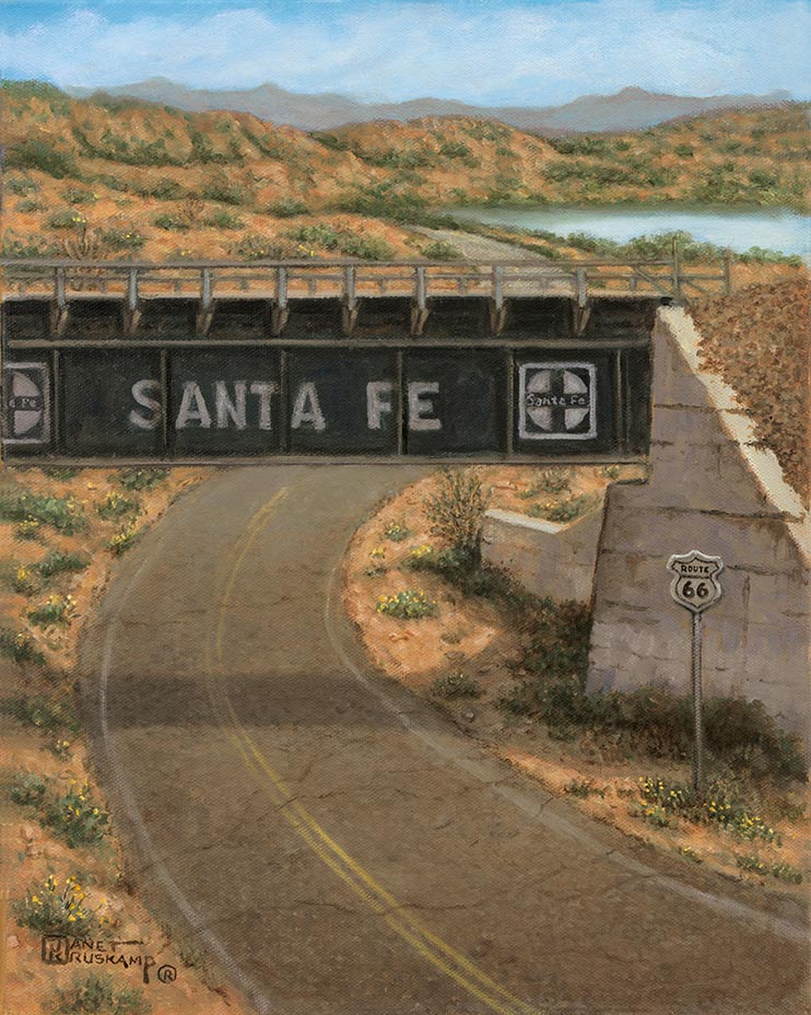 The dusty, curving desert highway runs under the green Santa Fe Railway overcrossing. A small white Route 66 sign stands on the roadside next to the concrete abutment. The hills in the background foreshadow the taller mountains at the horizon. The light blue sky is reflected in a body of water off the the right in the background.