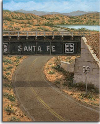 The dusty, curving desert highway runs under the green Santa Fe Railway overcrossing. A small white Route 66 sign stands on the roadside next to the concrete abutment. The hills in the background foreshadow the taller mountains at the horizon. The light blue sky is reflected in a body of water off the the right in the background.